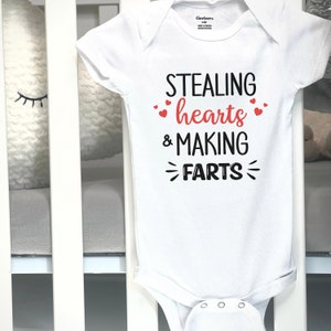 May include: White baby onesie with the text "STEALING hearts & MAKING FARTS" in black and red. The onesie is hanging on a white crib. The text is in a playful font with heart accents.