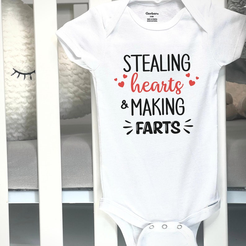 May include: White baby onesie with the text "STEALING hearts & MAKING FARTS" in black and red. The onesie is hanging on a white crib. The text is in a playful font with heart accents.