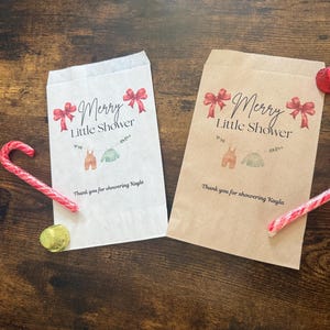May include: Two paper gift bags with the text "Merry Little Shower" and baby clothes illustrations. Red bows and candy canes decorate the scene. One bag is white, the other is brown. A gold foil-wrapped chocolate coin is also present.