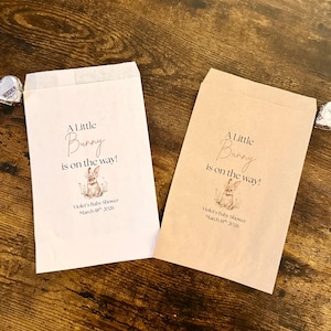 May include: Two paper favor bags with the text "A Little Bunny is on the way!" and a bunny illustration. The bags are for Violet's Baby Shower, March 18th, 2026. One bag is light pink, the other is tan.