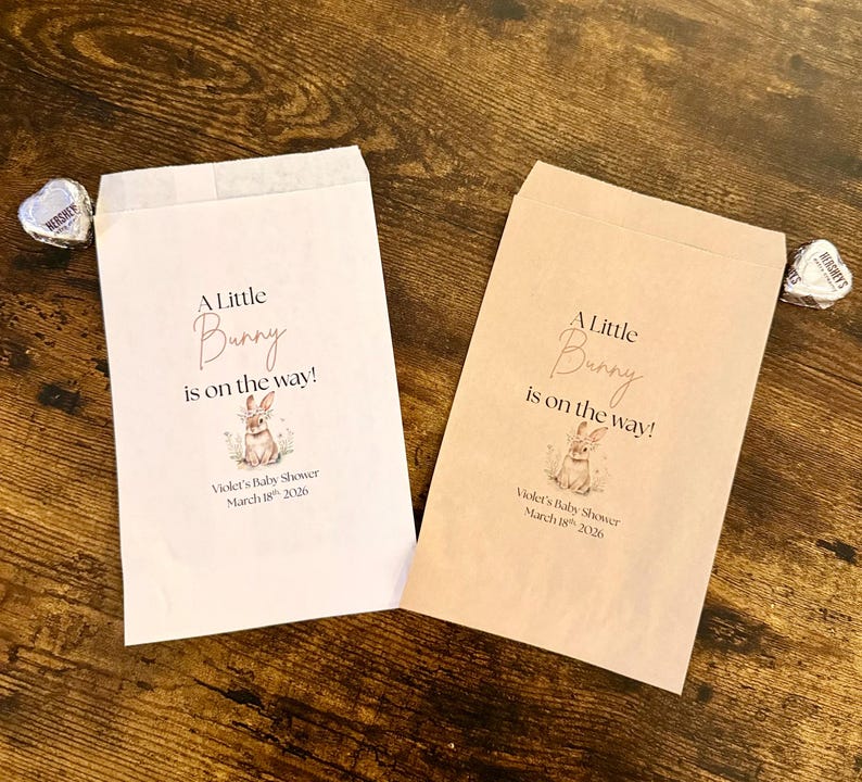 May include: Two paper favor bags with the text "A Little Bunny is on the way!" and a bunny illustration. The bags are for Violet's Baby Shower, March 18th, 2026. One bag is light pink, the other is tan.