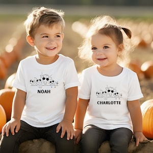 May include: Two children wearing white t-shirts with a black and white graphic of farm animals wearing sunglasses. The shirts have the names "NOAH" and "CHARLOTTE" printed below the graphic. The background is a pumpkin patch.