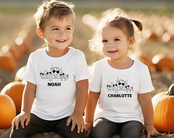 May include: Two children wearing white t-shirts featuring a black and white graphic of farm animals in sunglasses. The shirts have the names "NOAH" and "CHARLOTTE" printed below the graphic. The background is a pumpkin patch.