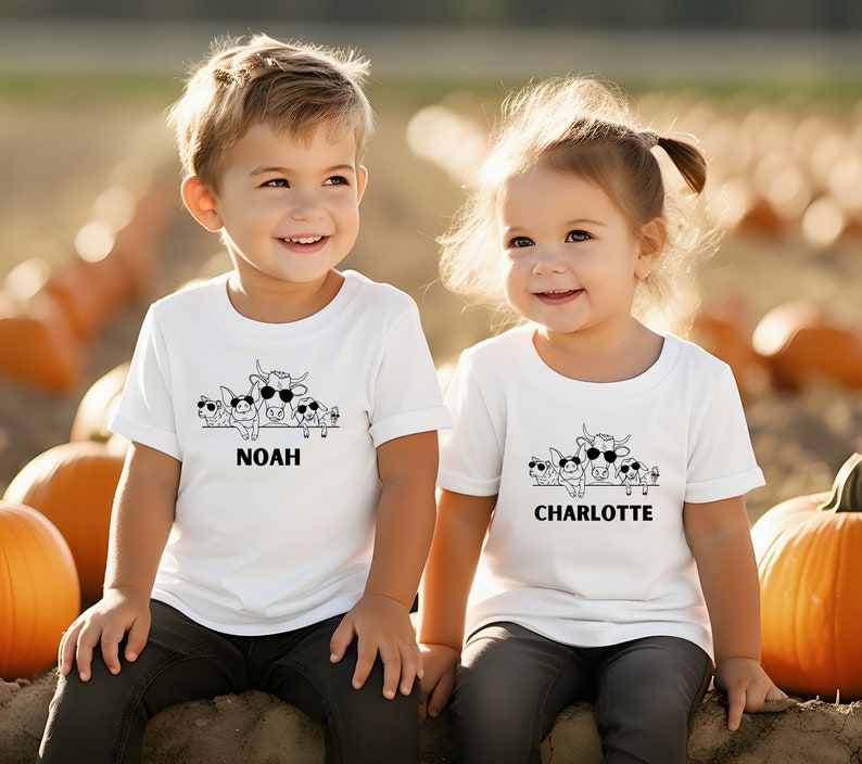 Puede incluir: Dos ni&ntilde;os con camisetas blancas con un gr&aacute;fico en blanco y negro de animales de granja con gafas de sol. Las camisetas tienen los nombres "NOAH" y "CHARLOTTE" impresos debajo del gr&aacute;fico. El fondo es un campo de calabazas.