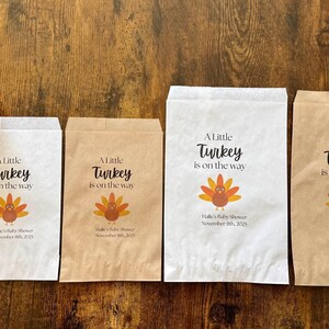May include: Four paper bags with the text "A Little Turkey is on the way" and a turkey graphic. Two pumpkins are on the wooden surface. The bags are white and brown, with the baby shower date printed on them.