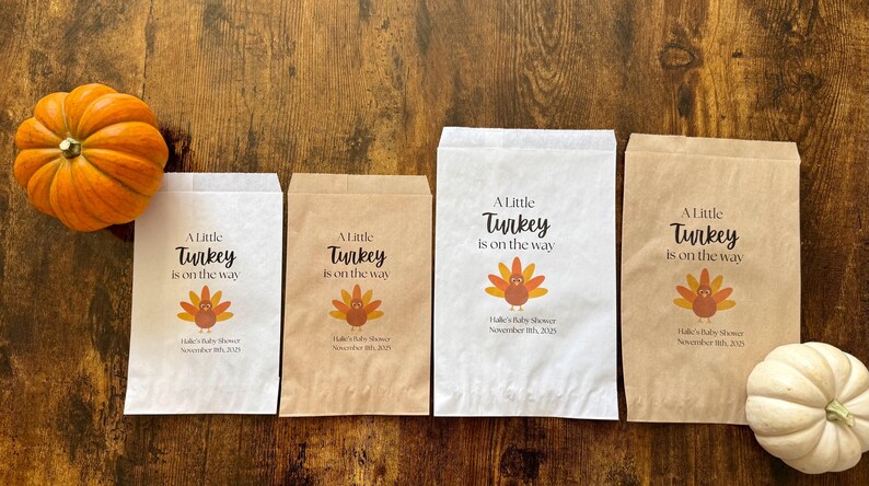 May include: Four paper bags with the text "A Little Turkey is on the way" and a turkey graphic. Two pumpkins are on the wooden surface. The bags are white and brown, with the baby shower date printed on them.
