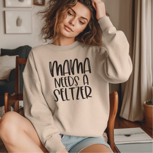 Puede incluir: Sudadera beige con el texto negro "MAMA NEEDS A SELTZER". La sudadera tiene mangas largas y cuello redondo. La imagen muestra a una persona usando la sudadera.