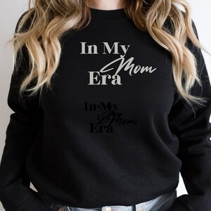 May include: Black sweatshirt with the text "In My Era Mom" in a white, cursive font. Below, a smaller, darker version of the text is visible. The sweatshirt is a casual, everyday garment.