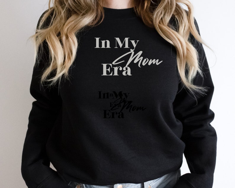 May include: Black sweatshirt with the text "In My Era Mom" in a white, cursive font. Below, a smaller, darker version of the text is visible. The sweatshirt is a casual, everyday garment.
