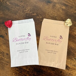 May include: Two paper favour bags, one white and one tan, with the text "A Little Butterfly Is On The Way!" in pink script. Each bag has a small butterfly graphic and the baby shower date. A heart-shaped sweet is attached to each bag.