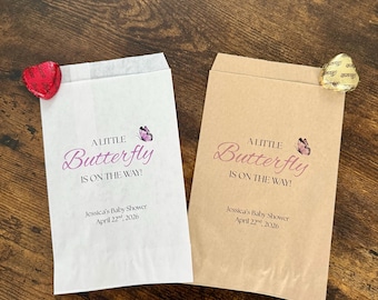 Puede incluir: Dos bolsas de papel para regalos, una blanca y otra beige, con el texto "A Little Butterfly Is On The Way!" en escritura rosa. Cada bolsa tiene un pequeño gráfico de mariposa y la fecha del baby shower. Un caramelo en forma de corazón está adjunto a cada bolsa.