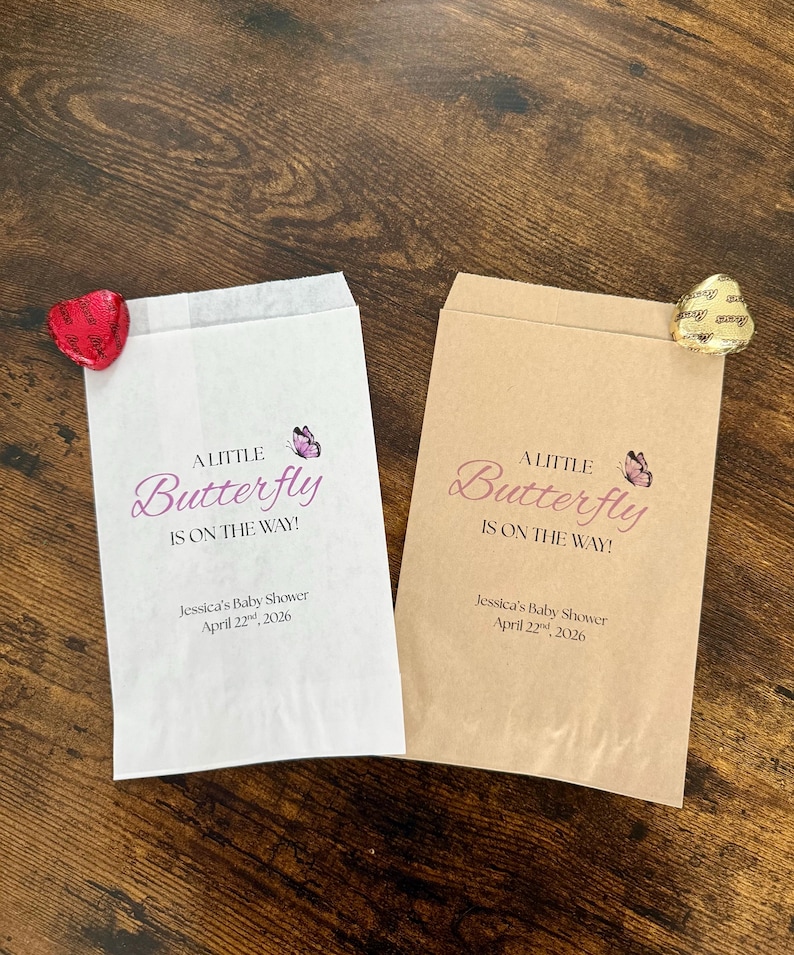 May include: Two paper favour bags, one white and one tan, with the text "A Little Butterfly Is On The Way!" in pink script. Each bag has a small butterfly graphic and the baby shower date. A heart-shaped sweet is attached to each bag.