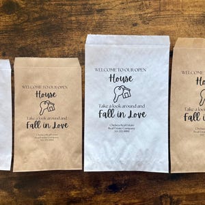 May include: Four paper bags with the text "WELCOME TO OUR OPEN House Take a look around and Fall in Love". The bags are white and brown, with a key and house graphic.