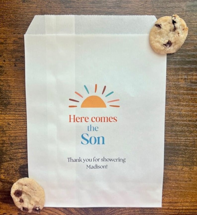 以下が含まれることがあります： 「Here comes the Son」の文字とオレンジ、ティール、赤色の太陽のグラフィックが入った白い紙袋。バッグには「Thank you for showering Madison!」の文字も含まれています。チョコレートチップクッキーが2つ、バッグの隣にあります。