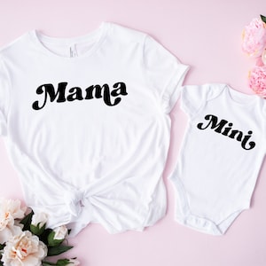 May include: White t-shirt with the word "Mama" in a black retro font, next to a white baby onesie with the word "Mini" in a matching font. The items are on a pink surface with floral accents.
