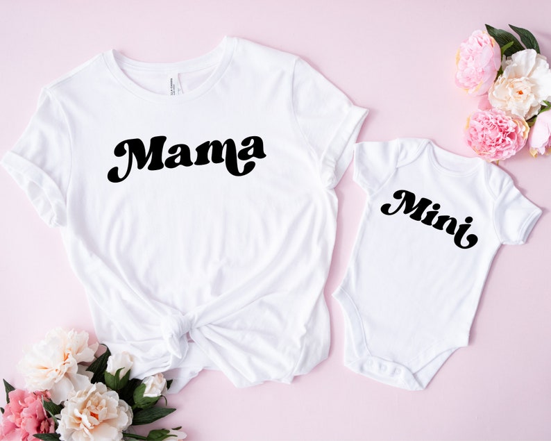 May include: White t-shirt with the word "Mama" in a black retro font, next to a white baby onesie with the word "Mini" in a matching font. The items are on a pink surface with floral accents.