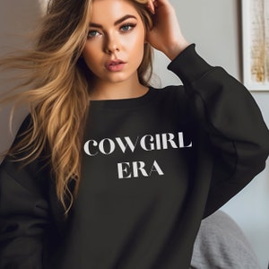 May include: Black sweatshirt with the text "COWGIRL ERA" in white, bold, capital letters. The sweatshirt has a crew neck and long sleeves. The model in the image has long blonde hair and is looking directly at the camera.