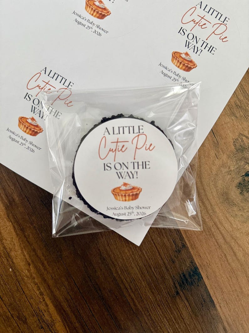 Op de afbeelding: Een ronde sticker in een doorzichtig plastic hoesje met de tekst A LITTLE Cutie Pie IS ON THE WAY!, een illustratie van een kleine taart en de tekst Jessica's Baby Shower August 25th, 2026. Een vel met identieke stickers is erachter zichtbaar.