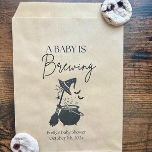 May include: A light brown paper bag with the words "A Baby Is Brewing" and a Halloween-themed illustration. The illustration features a witch's hat, broom, cauldron, and bats. The bag also includes the text "Emily's Baby Shower October 7th, 2024". Two biscuits are on the bag.