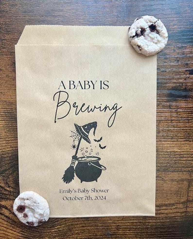 May include: A light brown paper bag with the words "A Baby Is Brewing" and a Halloween-themed illustration. The illustration features a witch's hat, broom, cauldron, and bats. The bag also includes the text "Emily's Baby Shower October 7th, 2024". Two biscuits are on the bag.