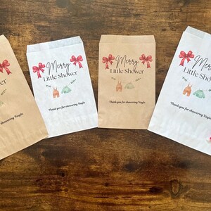 May include: Four paper gift bags with the text "Merry Little Shower" and illustrations of baby clothes. The bags are in shades of brown and white, with red bows and candy canes. A yellow heart and a red ornament are also present.