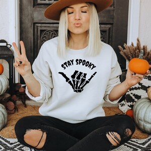 Może przedstawiać: Kremowa bluza z napisem "STAY SPOOKY" i wzorem szkieletu dłoni. Na zdjęciu dynie i jesienne dekoracje, tworzące motyw Halloween.