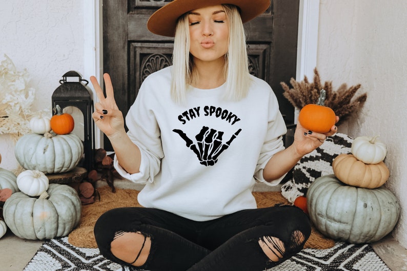 Może przedstawiać: Kremowa bluza z napisem "STAY SPOOKY" i wzorem szkieletu dłoni. Na zdjęciu dynie i jesienne dekoracje, tworzące motyw Halloween.