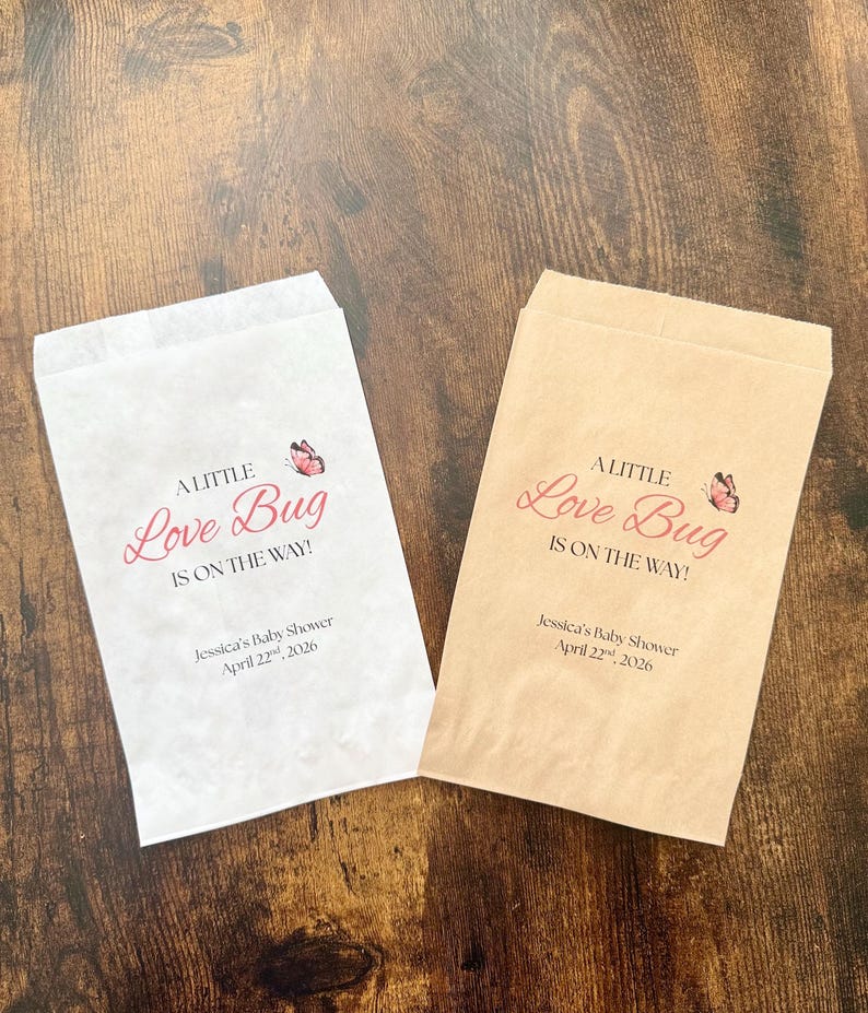 May include: Two paper favor bags, one white and one tan, with the text "A Little Love Bug Is On The Way!" in pink script. A pink butterfly graphic is above the text. The bags are for Jessica's Baby Shower, April 22, 2026.