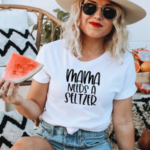 May include: White t-shirt with the text "MAMA NEEDS A SELTZER" in black script. The person is holding a watermelon slice. The background includes a wicker chair, decorative pillows, and a bowl of fruit.