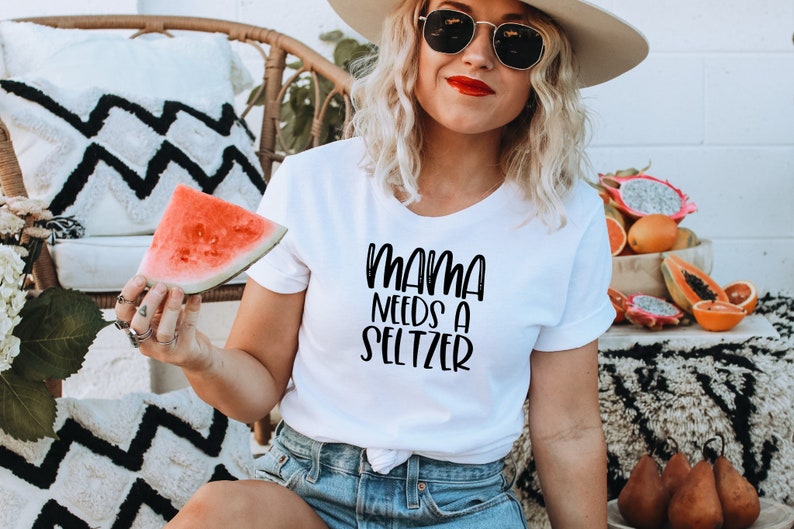 May include: White t-shirt with the text "MAMA NEEDS A SELTZER" in black script. The person is holding a watermelon slice. The background includes a wicker chair, decorative pillows, and a bowl of fruit.