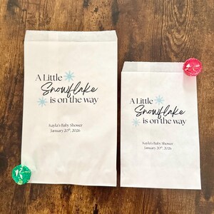 May include: Two white paper favor bags with the text "A Little Snowflake is on the way" in black and light blue. The bags are for Kayla's Baby Shower, January 20th, 2026. One bag has a green candy, the other has a pink candy.