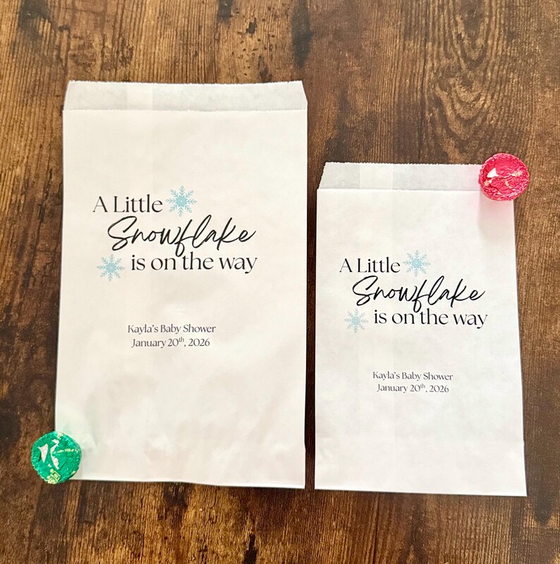 May include: Two white paper favor bags with the text "A Little Snowflake is on the way" in black and light blue. The bags are for Kayla's Baby Shower, January 20th, 2026. One bag has a green candy, the other has a pink candy.