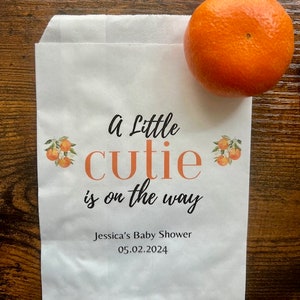 K&ouml;nnte beinhalten: Eine wei&szlig;e Papiert&uuml;te mit dem Text "A Little cutie is on the way" in Schwarz und Orange. Die T&uuml;te hat Orangenillustrationen und den Text "Jessica's Baby Shower 05.02.2024". Eine Orange liegt auf der T&uuml;te.
