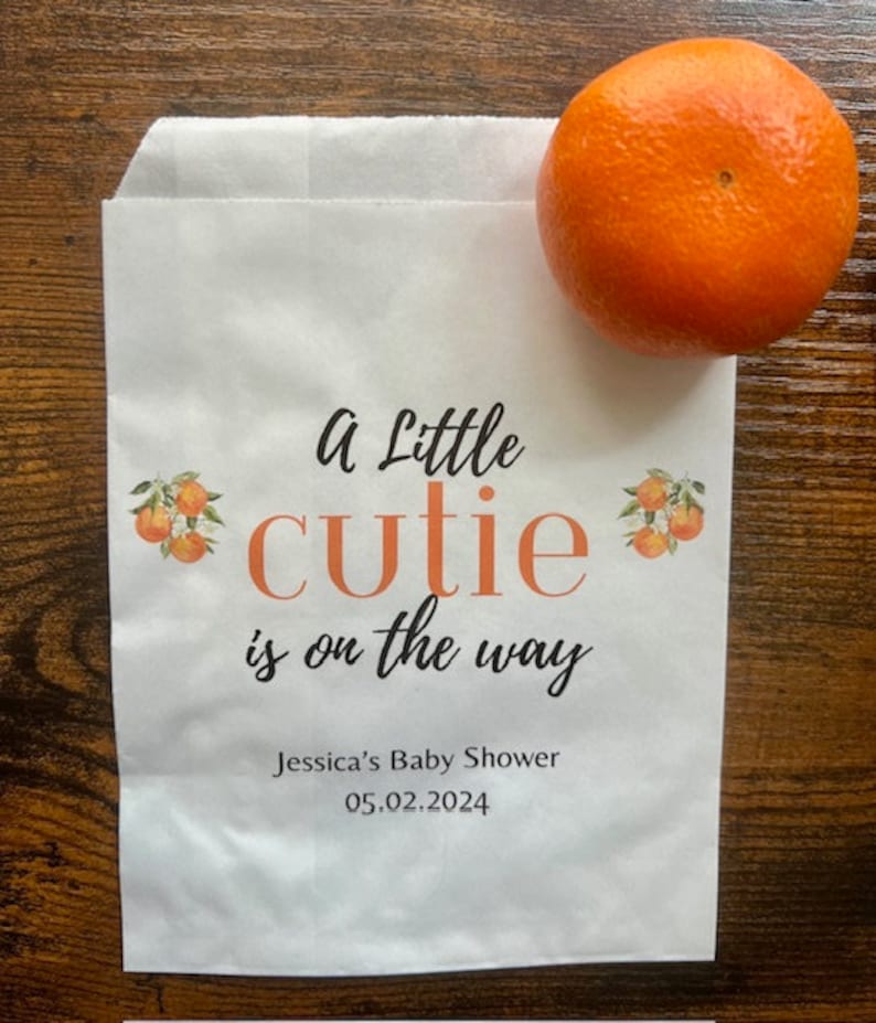 K&ouml;nnte beinhalten: Eine wei&szlig;e Papiert&uuml;te mit dem Text "A Little cutie is on the way" in Schwarz und Orange. Die T&uuml;te hat Orangenillustrationen und den Text "Jessica's Baby Shower 05.02.2024". Eine Orange liegt auf der T&uuml;te.