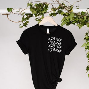 Może przedstawiać: Czarny t-shirt z napisem "Philly" ułożonym czterokrotnie w białym skrypcie. Koszulka wisi na drewnianym wieszaku na tle zieleni. Rękawy są podwinięte.