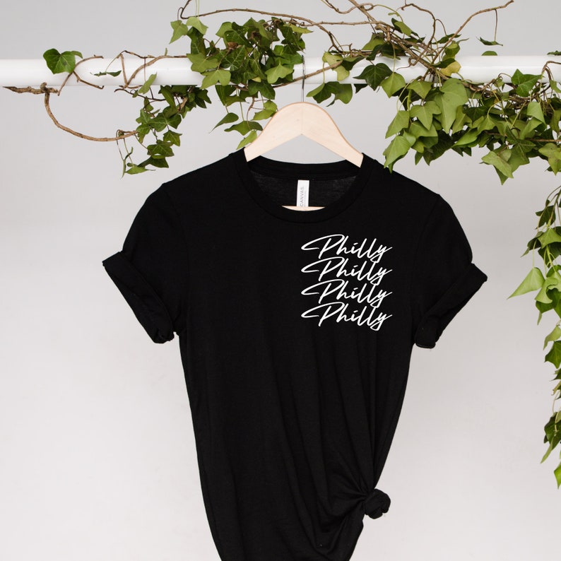 Może przedstawiać: Czarny t-shirt z napisem "Philly" ułożonym czterokrotnie w białym skrypcie. Koszulka wisi na drewnianym wieszaku na tle zieleni. Rękawy są podwinięte.