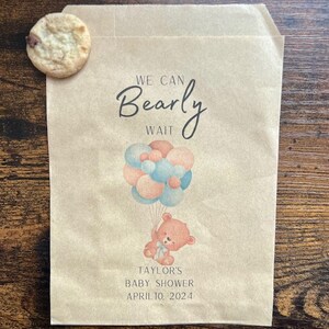 May include: A brown paper bag with a cookie and a baby shower design. The bag features a teddy bear holding balloons and the text "We Can Bearly Wait." The baby shower is for Taylor and is scheduled for April 10, 2024.