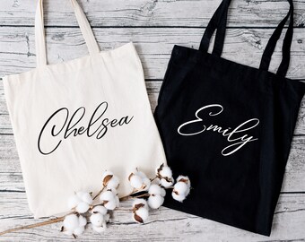 Peut inclure: Deux sacs fourre-tout, un crème et un noir, avec les noms "Chelsea" et "Emily" en écriture cursive élégante. Les sacs ont de longues anses et sont faits d'un tissu robuste. Une branche de coton ajoute une touche décorative.