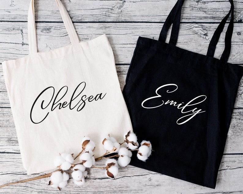 May include: Two tote bags, one cream and one black, with the names "Chelsea" and "Emily" in elegant cursive script. The bags have long handles and are made of a sturdy fabric. A sprig of cotton adds a decorative touch.