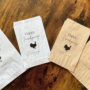 May include: Four paper treat bags with the text "Happy Friendsgiving!" and a turkey silhouette. Two bags are white, and two are tan. The bags are on a wooden surface.
