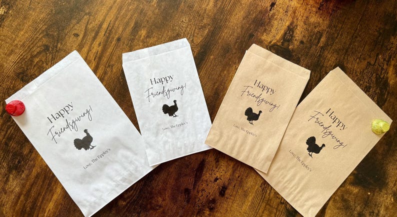 May include: Four paper treat bags with the text "Happy Friendsgiving!" and a turkey silhouette. Two bags are white, and two are tan. The bags are on a wooden surface.