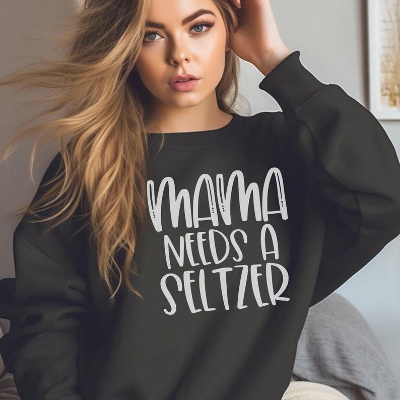 May include: Black sweatshirt with the white text "MAMA NEEDS A SELTZER". The sweatshirt has a crew neck and long sleeves. The text is in a handwritten style font.