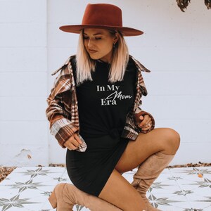 May include: A woman wearing a brown fedora, a plaid shirt, a black dress, and tan boots. The black t-shirt has the text "In My Mom Era" in white script. The outfit is styled with a casual, yet fashionable aesthetic.