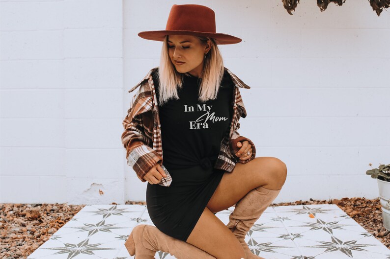May include: A woman wearing a brown fedora, a plaid shirt, a black dress, and tan boots. The black t-shirt has the text "In My Mom Era" in white script. The outfit is styled with a casual, yet fashionable aesthetic.