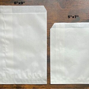 May include: Two white paper bags, one measuring 6" x 9" and the other 5" x 7". The bags are rectangular with a folded top. The paper has a slightly textured appearance. The bags are likely for packaging or storage.