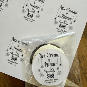 May include: Round white adhesive labels featuring black text that reads We Created a Monster, a graphic of a baby carriage, spider webs, and stars. Below the carriage, the text reads Jessica's Baby Shower October 31, 2026. One label is on a cookie.