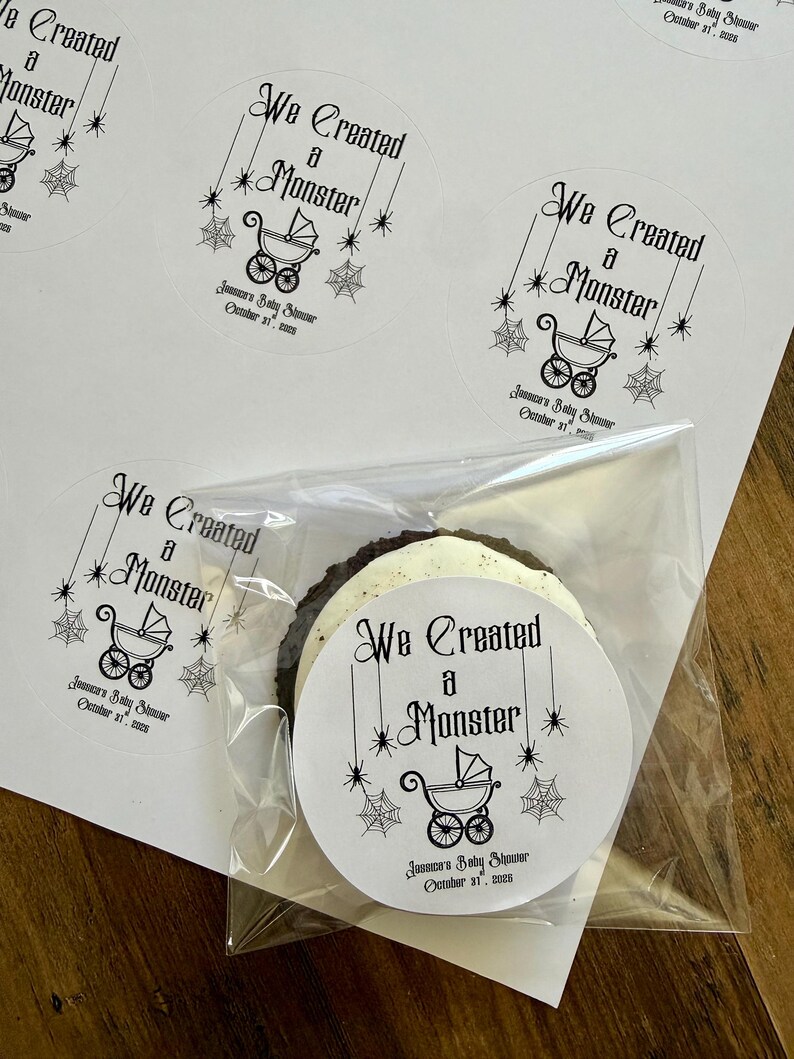 May include: Round white adhesive labels featuring black text that reads We Created a Monster, a graphic of a baby carriage, spider webs, and stars. Below the carriage, the text reads Jessica's Baby Shower October 31, 2026. One label is on a cookie.