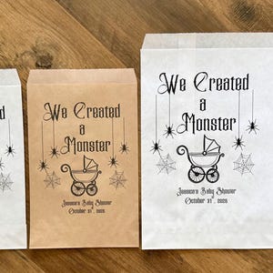 May include: Four rectangular paper favor bags in white and brown, featuring the black printed text We Created a Monster, a baby carriage icon, spider webs, and spiders, with the text Jameson's Baby Shower October 31, 2024 below the carriage.