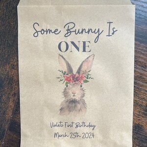以下が含まれることがあります： 「Some Bunny Is ONE」の文字と、花の冠をつけたウサギの水彩イラストが描かれた茶色の紙袋。「Violet's First Birthday March 25th 2024」の文字も印刷されています。