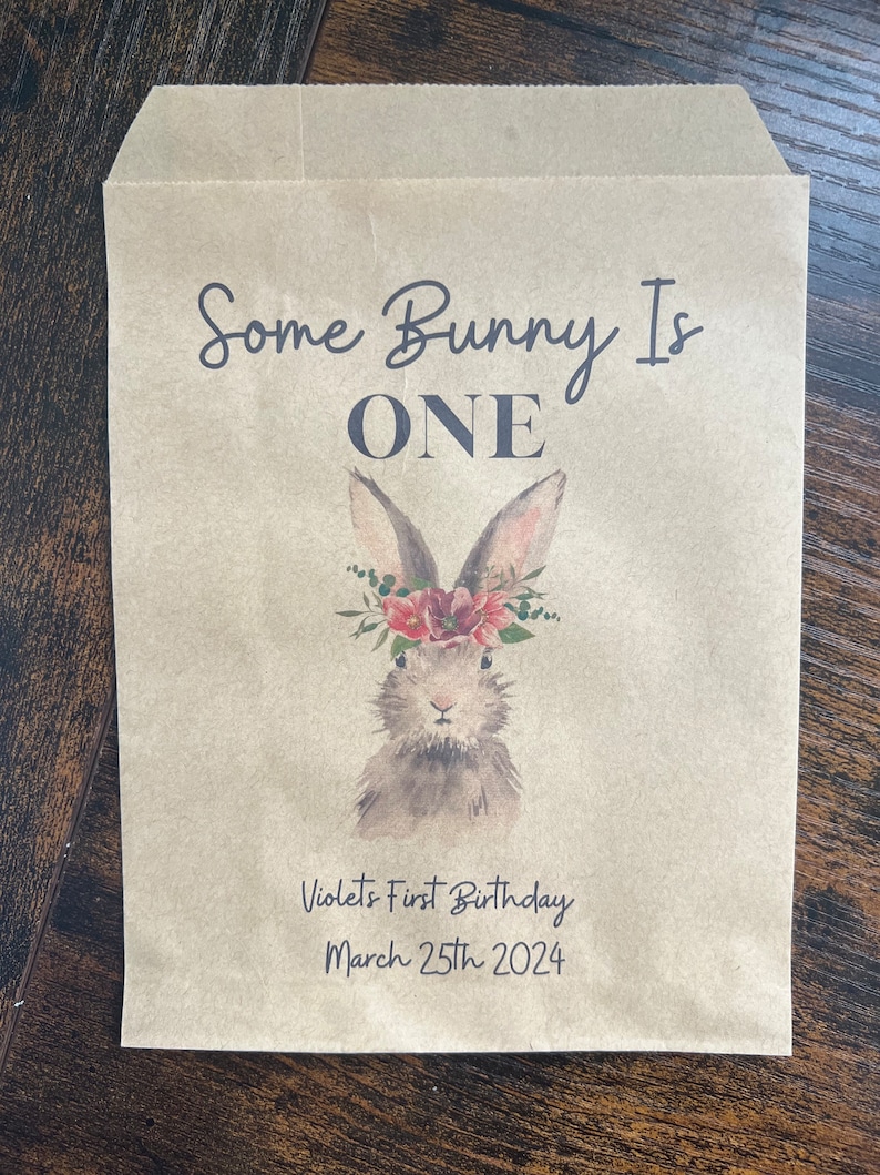 以下が含まれることがあります： 「Some Bunny Is ONE」の文字と、花の冠をつけたウサギの水彩イラストが描かれた茶色の紙袋。「Violet's First Birthday March 25th 2024」の文字も印刷されています。